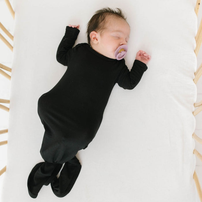 A sleeping infant in a black knotted gown with fold-over mittens and a tie bottom for diaper changes, lying on a white surface.