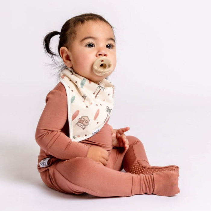 A baby sitting and wearing a red bamboo romper with a patterned bib.