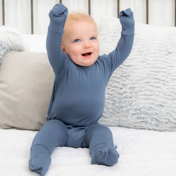 A baby wearing a gray bamboo romper with long sleeves and footies, sitting on a soft surface with raised hands.